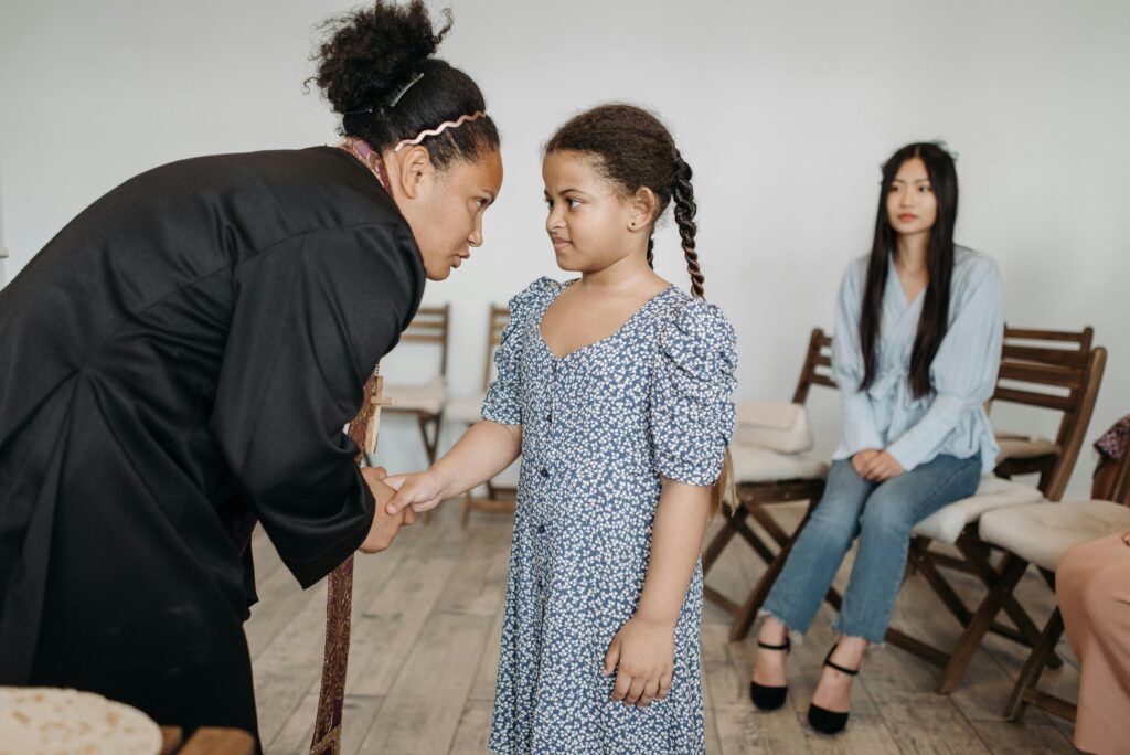 A clergy member interacts warmly with a young girl while others look on in a meeting room setting.